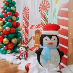 A penguin with santa hat and scarf next to christmas tree.