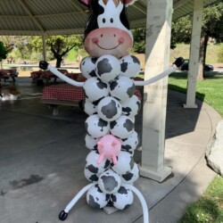 A cow balloon sculpture is standing in the middle of a park.