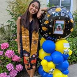 A woman in graduation gown standing next to balloons.
